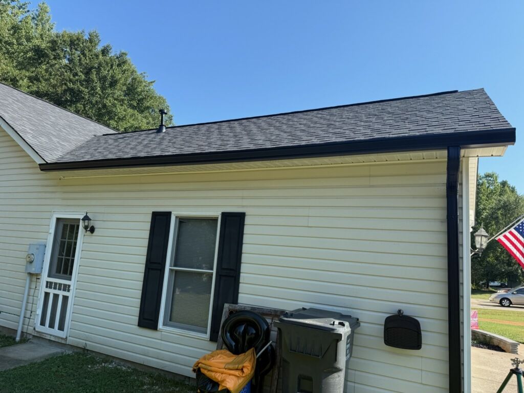 Side of a yellow sided house with a shingled roof and black seamless gutters and downspout.