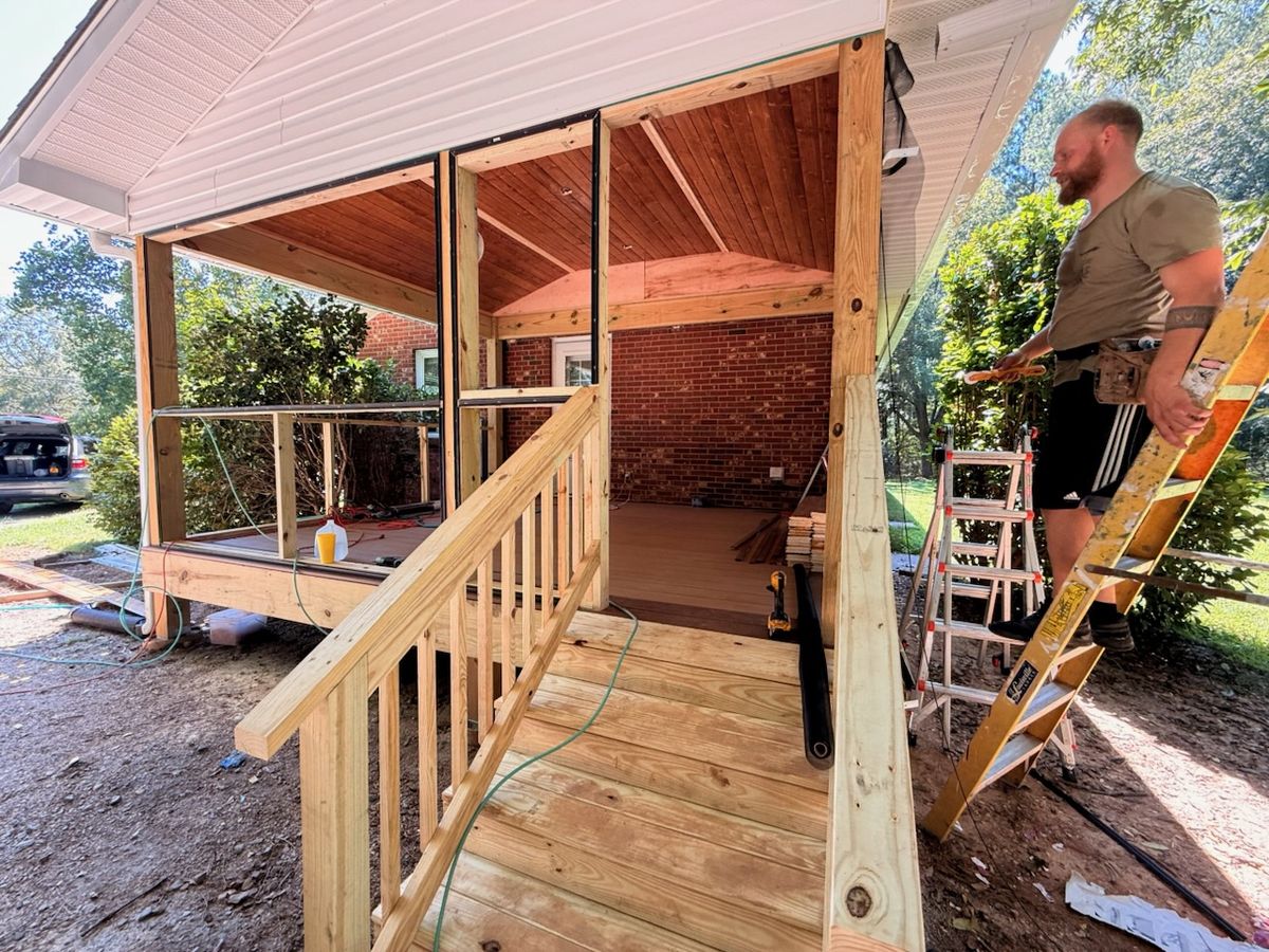 A worker on a ladder beside a new wooden ramp and covered porch with a vaulted wood ceiling on a brick house.
