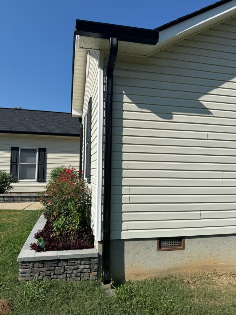 A black gutter downspout runs down the corner of a house with cream vinyl siding and a raised stone flower bed.