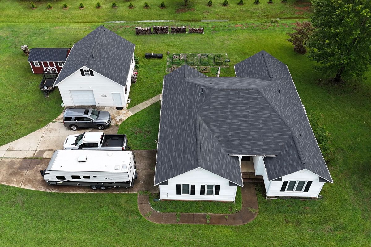 Aerial view of a white house and detached garage with dark gray asphalt shingle roofs, surrounded by green lawn.