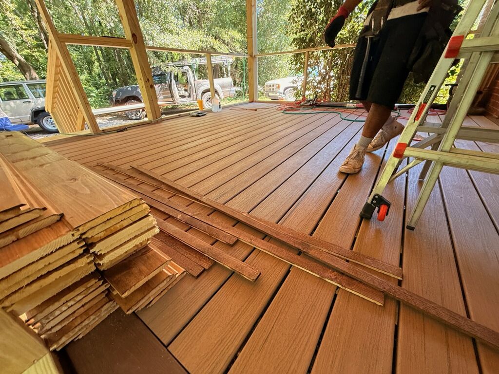A worker stands on a brown composite deck under construction, with stacked wood boards and a ladder nearby.